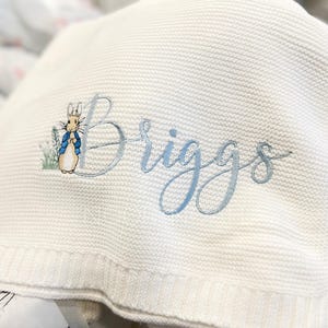 May include: White knitted baby blanket with the name "Briggs" embroidered in light blue script. A Peter Rabbit illustration is embroidered next to the first letter. The blanket has a textured appearance.