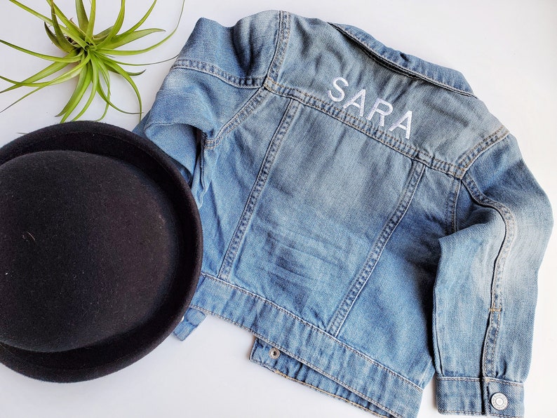 May include: A light blue denim jacket with white embroidery that says "SARA" on the back.