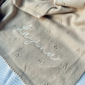 May include: A beige knitted baby blanket with the name "Layina" embroidered in white script. The blanket has a delicate openwork pattern and a scalloped edge. The brand name "NOOR+NOAH" is printed at the bottom.