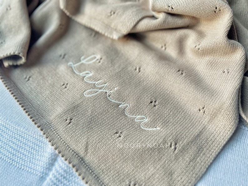 May include: A beige knitted baby blanket with the name "Layina" embroidered in white script. The blanket has a delicate openwork pattern and a scalloped edge. The brand name "NOOR+NOAH" is printed at the bottom.