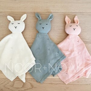 May include: Three bunny-shaped baby security blankets in white, blue, and pink. Each has a soft, textured body with a bunny head and embroidered facial features. The blankets are arranged on a light wood surface.