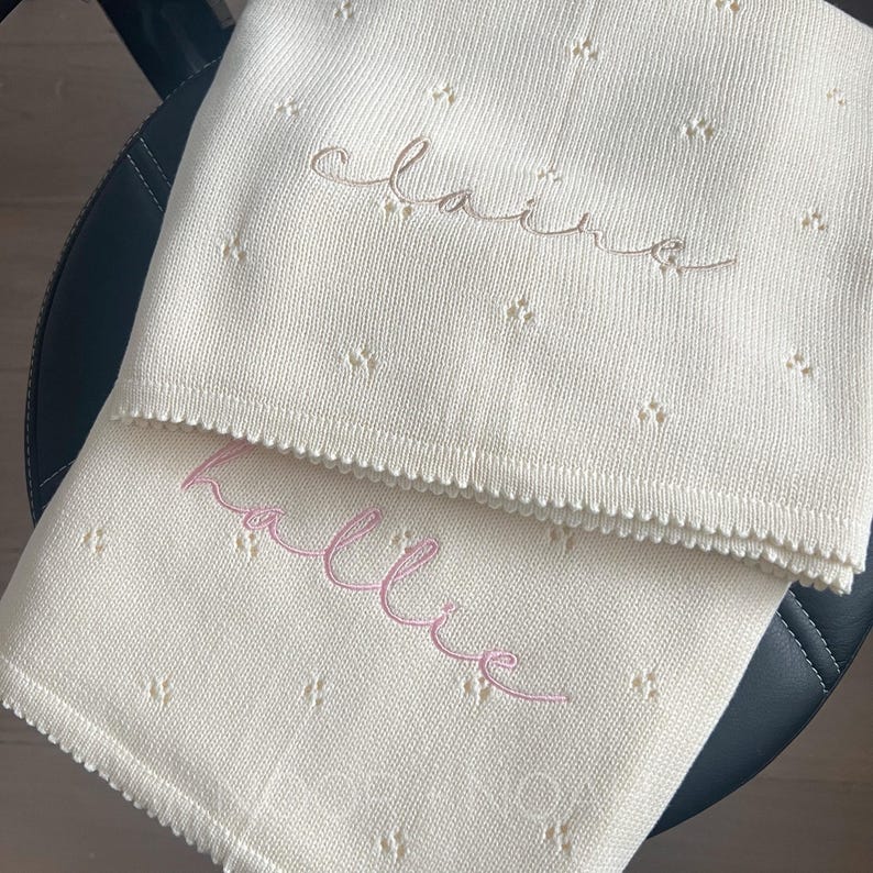 May include: Two cream-colored baby blankets with embroidered names. One blanket has the name "Claire" in a light brown script, and the other has the name "Hallie" in pink script. Both blankets have a delicate, dotted pointelle  pattern.