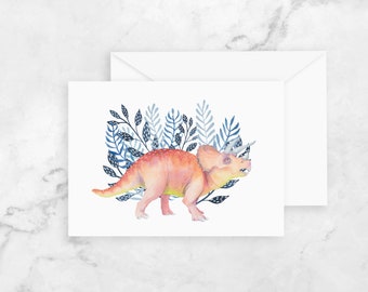 May include: Watercolor illustration of a Triceratops dinosaur on a white card. The dinosaur is peach and yellow, set against blue and navy foliage. A white envelope is visible behind the card. The image has a soft, artistic feel.