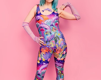 May include: A woman wearing a blue and purple one-piece jumpsuit with a colorful ocean-themed print. The jumpsuit features dolphins, sea turtles, jellyfish, and other marine life. She is wearing pink gloves and light pink boots.