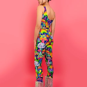 May include: A person wearing a colorful jumpsuit with a psychedelic mushroom and rainbow pattern. The jumpsuit has a scoop neck and thin straps. The person is wearing silver cowboy boots with fringe.