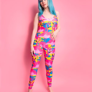 May include: A woman with blue hair is wearing a pink, yellow, and blue jumpsuit with a flamingo print. The jumpsuit has a scoop neck and a sleeveless design.