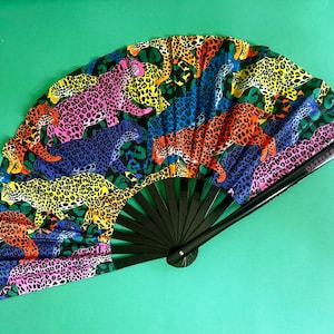 Peut inclure: Éventail pliable avec un cadre noir et un imprimé léopard coloré. Le tissu présente des léopards roses, jaunes, bleus et oranges sur fond noir. Le texte "DAZZLE WOW" est imprimé sur l'éventail, un accessoire de mode.