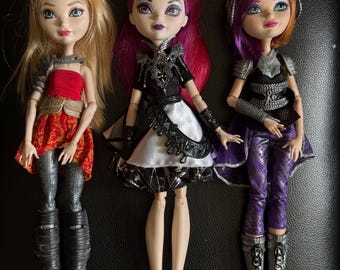 May include: Three fashion dolls are shown, each with unique outfits and accessories. The dolls have blonde, red, and purple hair. They are dressed in red, black, white, and purple clothing. The dolls are arranged on a black surface.