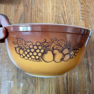 Könnte beinhalten: Eine braune und gelbe Glasschüssel mit einem schwarz-weißen Design aus Trauben und Äpfeln. Die Schüssel ist leer.