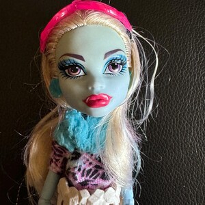 May include: A blue-skinned doll with blonde hair wearing a pink headband, a blue and white fuzzy scarf, and a pink and black patterned dress. The doll has white teeth around her waist.