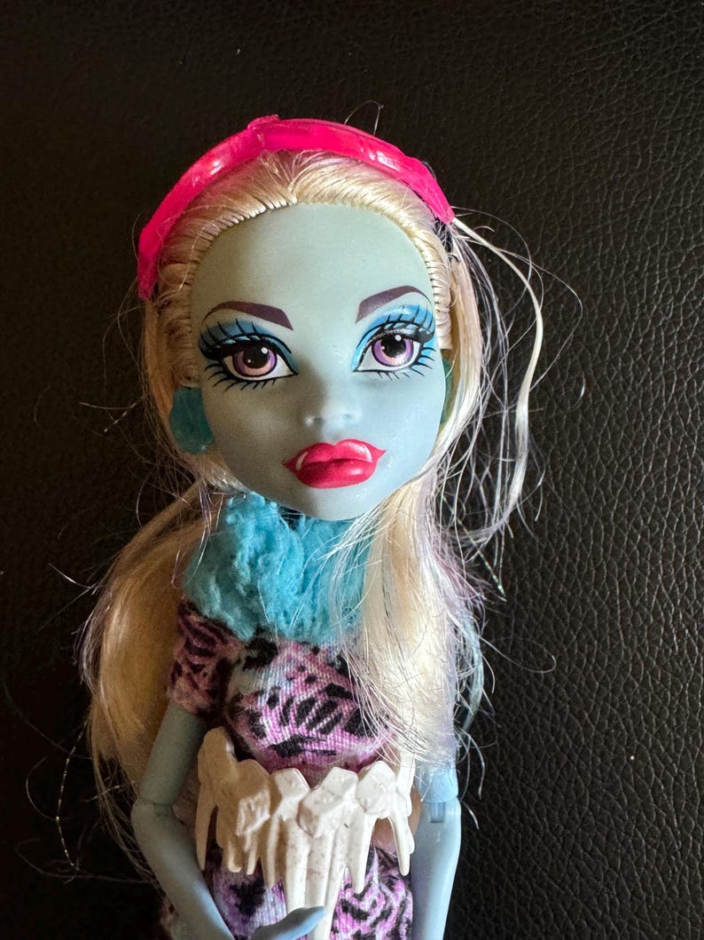 May include: A blue-skinned doll with blonde hair wearing a pink headband, a blue and white fuzzy scarf, and a pink and black patterned dress. The doll has white teeth around her waist.