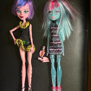 Könnte beinhalten: Zwei Monster High Puppen, eine mit lila und blauen Haaren und einem schwarzen und gelben Outfit, die andere mit blauen und rosa Haaren und einem blauen und schwarzen Kleid.