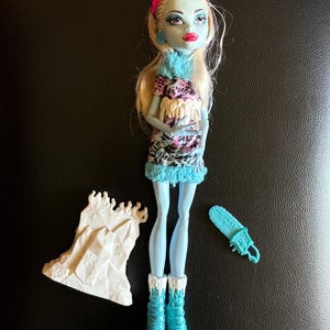 May include: A blue and white Monster High doll with blonde hair wearing a pink headband and a white and black patterned dress. The doll is holding a white ice sculpture and has a blue ice skate accessory.