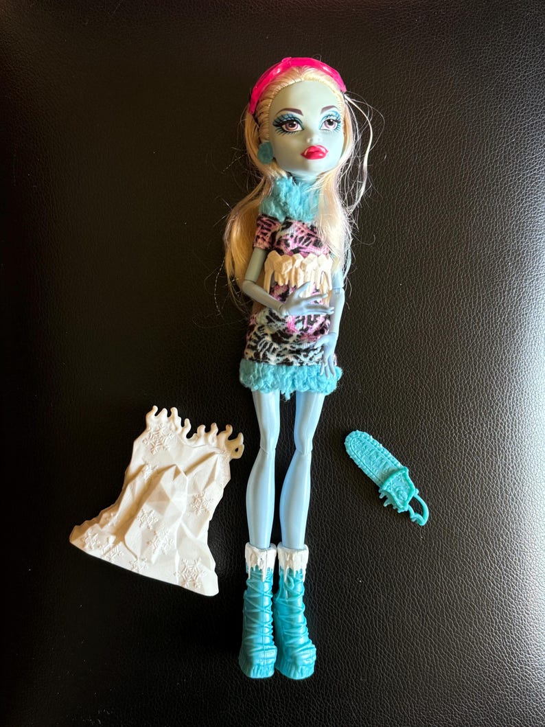 May include: A blue and white Monster High doll with blonde hair wearing a pink headband and a white and black patterned dress. The doll is holding a white ice sculpture and has a blue ice skate accessory.