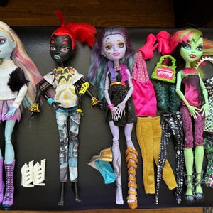 Peut inclure: Cinq poupées Monster High avec des coiffures et des tenues différentes. Les poupées portent des vêtements et des accessoires colorés, notamment des chaussures, des sacs à main et des bijoux. Les poupées sont posées dans différentes positions.