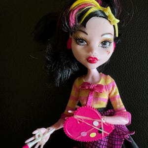 May include: A pink and black doll with yellow and pink hair. She is wearing a yellow and white striped shirt with a pink bow, a pink skirt with black spiderwebs, and a pink heart-shaped paint palette.