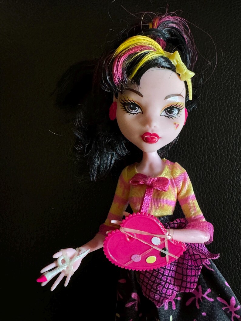 May include: A pink and black doll with yellow and pink hair. She is wearing a yellow and white striped shirt with a pink bow, a pink skirt with black spiderwebs, and a pink heart-shaped paint palette.