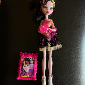 May include: A pink and black doll with black hair and yellow streaks. She is wearing a pink and yellow striped shirt, a black and white skirt with a pink heart, and white socks with pink shoes. She is holding a pink paint palette with a paintbrush. The doll is standing in front of a pink picture frame with a picture of a doll inside.