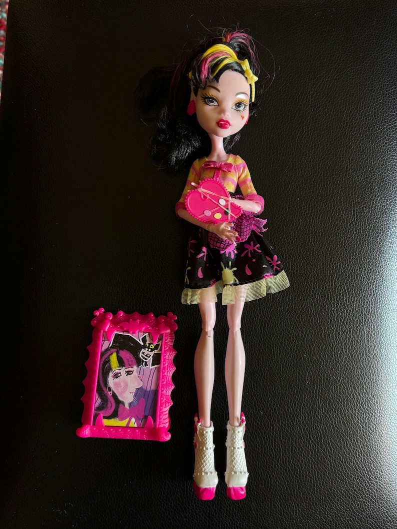 May include: A pink and black doll with black hair and yellow streaks. She is wearing a pink and yellow striped shirt, a black and white skirt with a pink heart, and white socks with pink shoes. She is holding a pink paint palette with a paintbrush. The doll is standing in front of a pink picture frame with a picture of a doll inside.