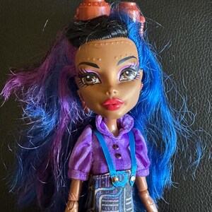 May include: A doll with blue and purple hair, brown skin, and brown eyes. She is wearing a purple shirt with blue straps and a black and blue skirt with a circuit board pattern. She has two brown gears on her head.
