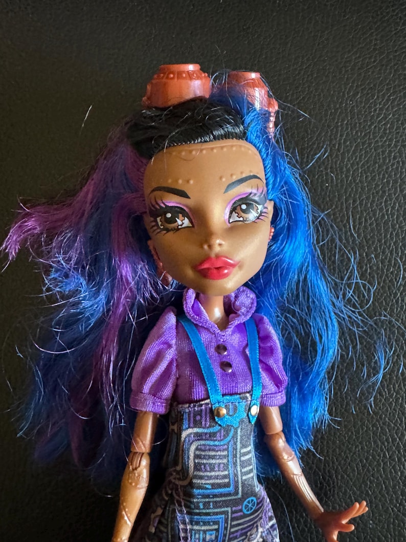 May include: A doll with blue and purple hair, brown skin, and brown eyes. She is wearing a purple shirt with blue straps and a black and blue skirt with a circuit board pattern. She has two brown gears on her head.