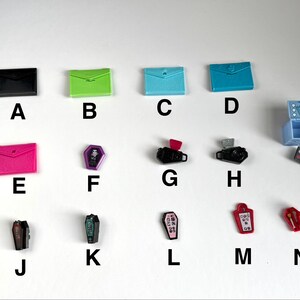 Op de afbeelding: Een verzameling miniatuurspeelgoed, waaronder kleurrijke enveloppen, doodskisten, een kaptafel en camera's. De items zijn in verschillende kleuren zoals zwart, groen, blauw, roze en rood. Het speelgoed is op een wit oppervlak geplaatst.