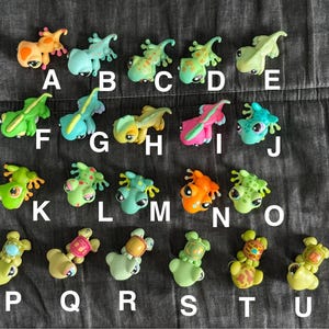 May include: A collection of 20 small plastic figurines of lizards and turtles in various colours, including green, blue, pink, and yellow. The figurines are arranged in two rows, with letters A through U labelling each figurine.