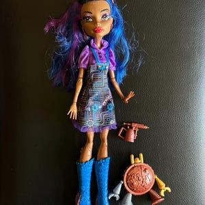 May include: A doll with blue and purple hair, brown skin, and a purple and black dress. She is wearing blue boots and holding a tool. The doll is part of a set that includes a robot with a shield and other tools.