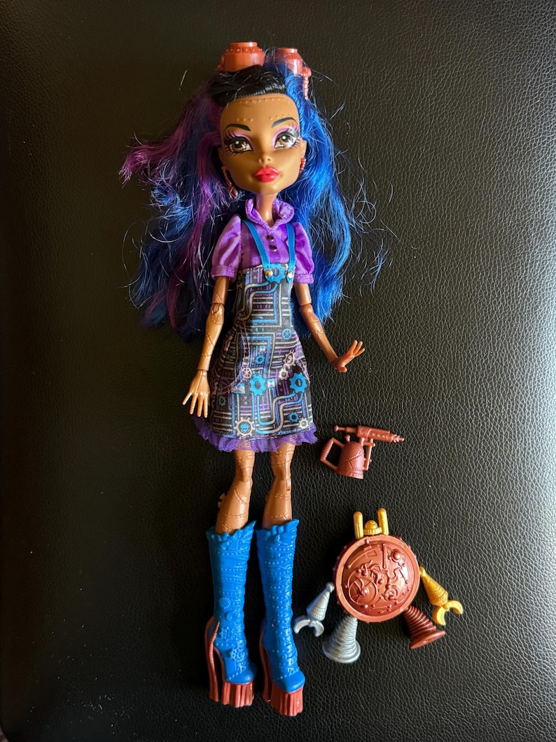 May include: A doll with blue and purple hair, brown skin, and a purple and black dress. She is wearing blue boots and holding a tool. The doll is part of a set that includes a robot with a shield and other tools.