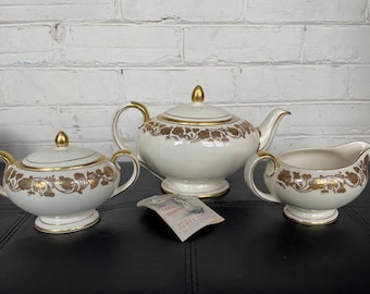 May include: A white teapot, creamer, and sugar bowl with gold floral designs. The teapot has a lid and a spout. The creamer and sugar bowl have lids. The set is on a black surface. The text on the tag reads "Tea & Teapot Made by James Beattie"