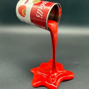 May include: A sculpture of a Campbell's tomato soup can with red liquid seemingly pouring out. The can is red and white with a tomato illustration. The liquid forms a star-shaped splash on the surface.