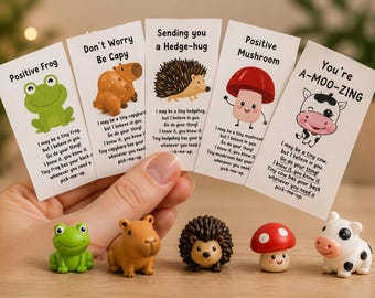 May include: A hand holds five cards with cartoon animal illustrations and positive messages. Below, matching figurines of a frog, capybara, hedgehog, mushroom, and cow are displayed. The cards read "Positive Frog", "Don't Worry Be Capy", "Sending you a Hedge-hug", "Positive Mushroom", and "You're A-MOO-ZING".