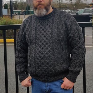 May include: A man wearing a gray cable knit sweater and blue jeans. He is standing in front of a black metal fence.