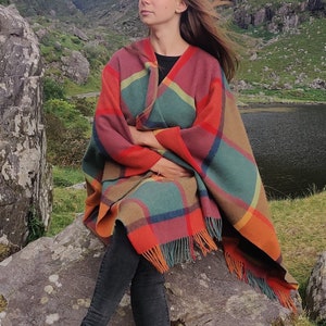 May include: A woman wearing black pants and black boots sits on a rock with a colorful plaid shawl wrapped around her shoulders. The shawl has a fringe border and is made of a soft, woven fabric.