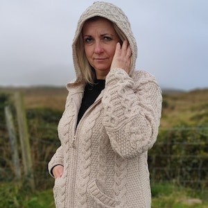 May include: A hooded, beige cable-knit sweater with a zipper closure and front pockets. The sweater has a textured pattern and is worn with black leggings. The background shows a road and grassy area.