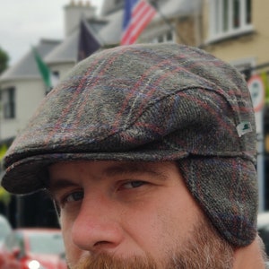 Può includere: Un uomo che indossa un cappello da newsboy in tweed grigio e rosso a quadri con le alette per le orecchie. Il cappello ha una fascia in pelle marrone e un logo verde. L'uomo indossa un maglione blu.