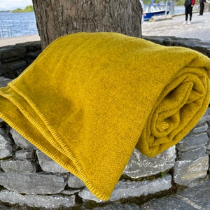 May include: A folded, mustard-yellow blanket rests on a stone structure near a body of water. The blanket is made of a textured material and has a rolled portion. The blanket is folded neatly, with a visible stitched edge.