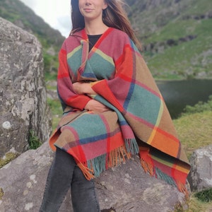 May include: A woman wearing a colorful plaid poncho with fringe, sitting on a large rock in a natural setting.