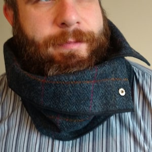 May include: A man with a beard is wearing a striped shirt and a dark blue and brown tweed neck warmer with a button on the side.