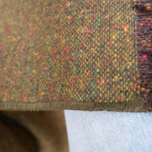 May include: Close-up of a brown tweed fabric with a green, yellow, and red flecked pattern. The fabric has a dark brown trim along the edge.