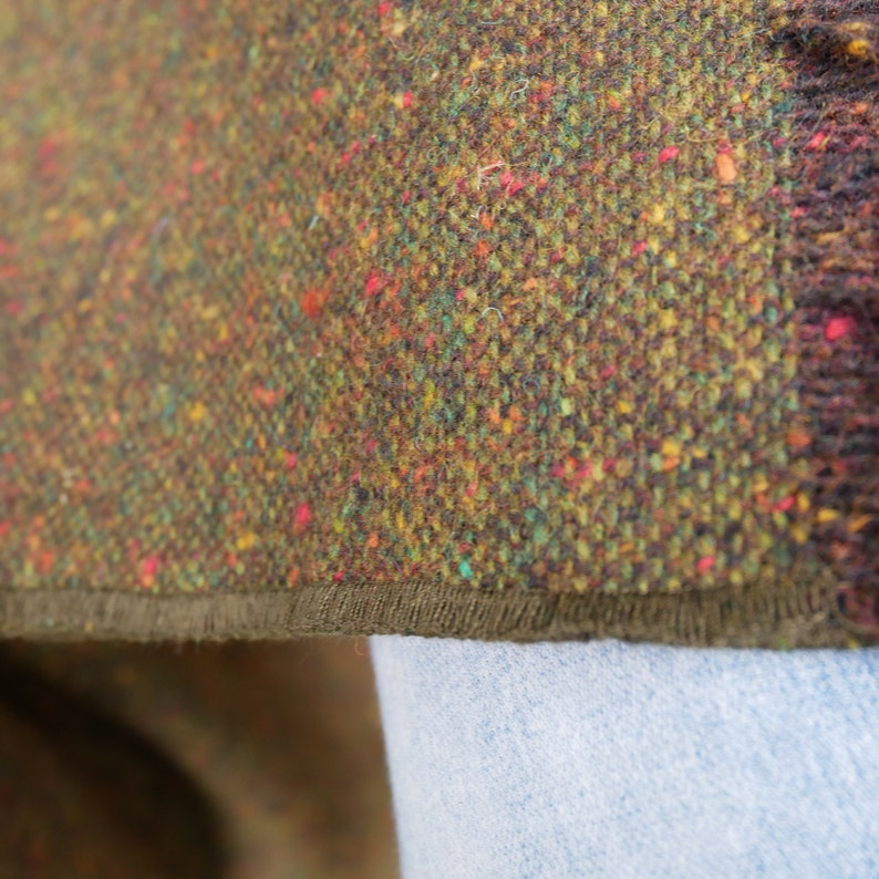 May include: Close-up of a brown tweed fabric with a green, yellow, and red flecked pattern. The fabric has a dark brown trim along the edge.