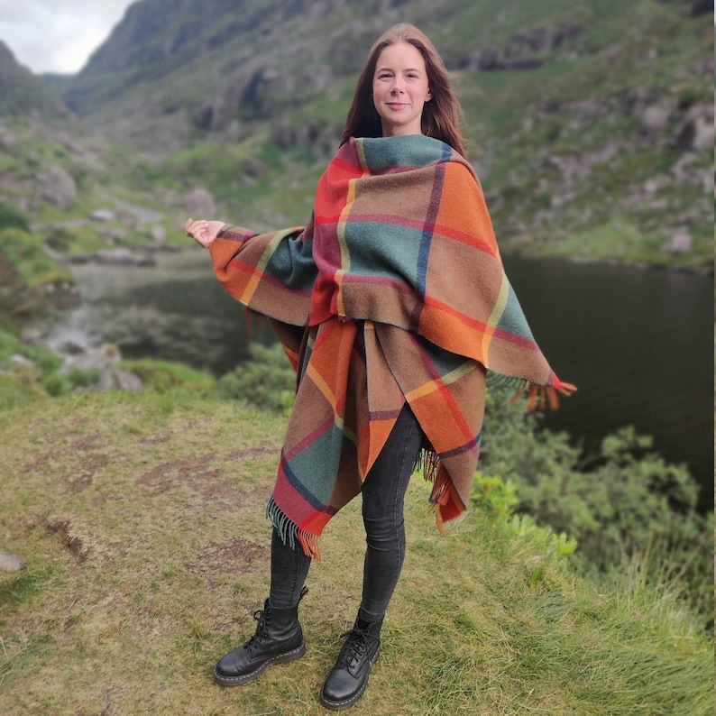 May include: A woman wearing a plaid poncho with fringe details. The poncho is in shades of brown, orange, green, and blue. She is standing in a grassy field with a lake in the background.