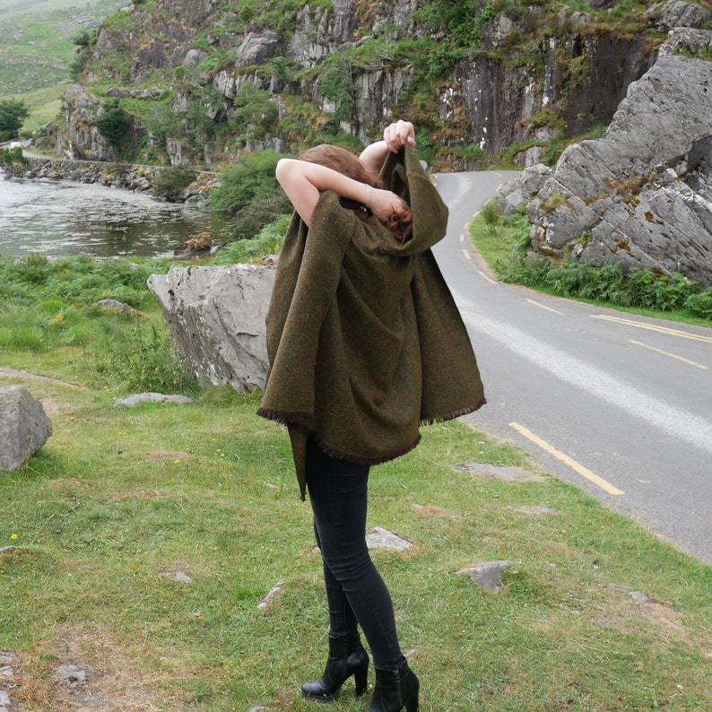 May include: A woman wearing a green hooded cape with a fringe trim, standing on a path with a rocky background.