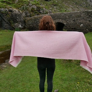 May include: A pink woven shawl with white fringe, held out in front of a person standing in a grassy field with a stone bridge in the background.
