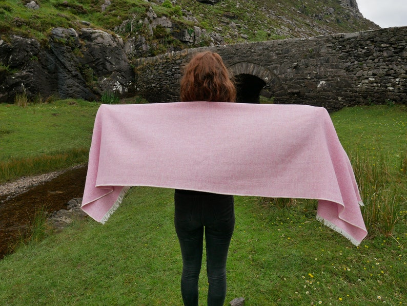 May include: A pink woven shawl with white fringe, held out in front of a person standing in a grassy field with a stone bridge in the background.