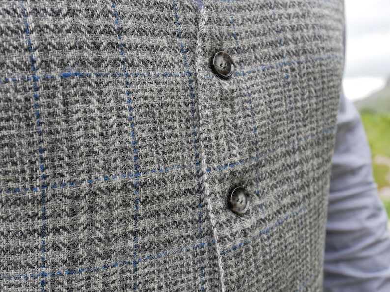 May include: A close-up of a gray and blue plaid vest with two black buttons. The vest is made of a soft, textured fabric.
