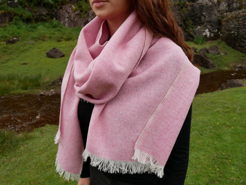 May include: A pink wool scarf with a white fringe. The scarf is folded and draped around a person's neck.