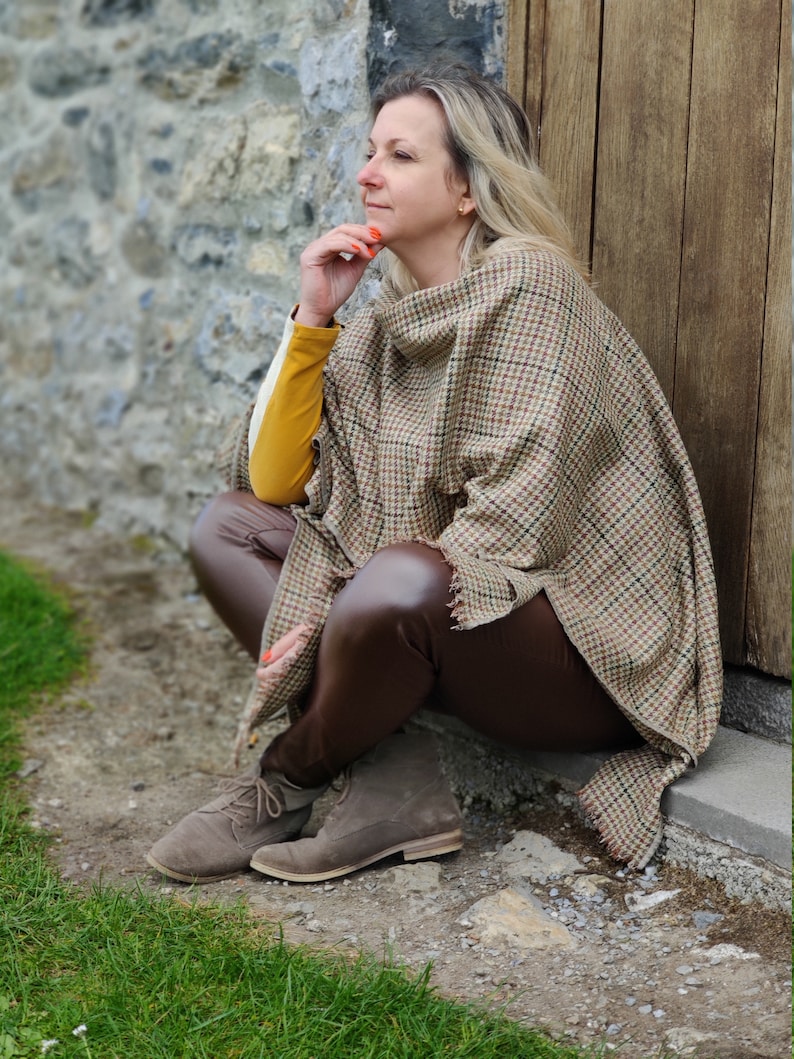 May include: A woman wearing a brown and white plaid poncho with a brown leather jacket and brown leather boots. She is sitting on a stone step with a stone wall behind her.