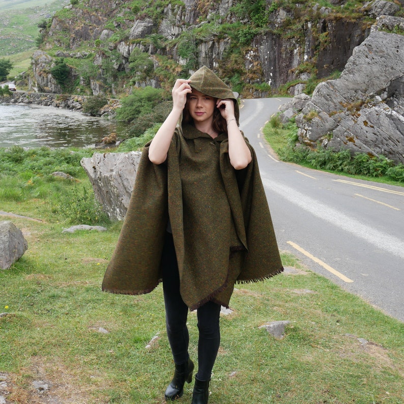 May include: A woman wearing a green hooded wool cape with a brown trim. The cape is worn over black pants and the woman is standing on a path with green grass and rocks.
