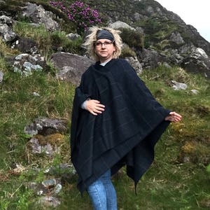 May include: A woman wearing a dark blue and black plaid poncho with a brown headband and jeans, standing in a grassy area with rocks and a mountain in the background.